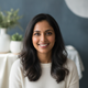 Smiling South Asian woman in her late 20s or early 30s, looking at the camera with a friendly expression. The background is a
